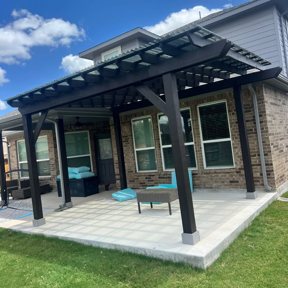Dark timber pergola with light-transmitting roof panels over a concrete patio slab beside a brick home.