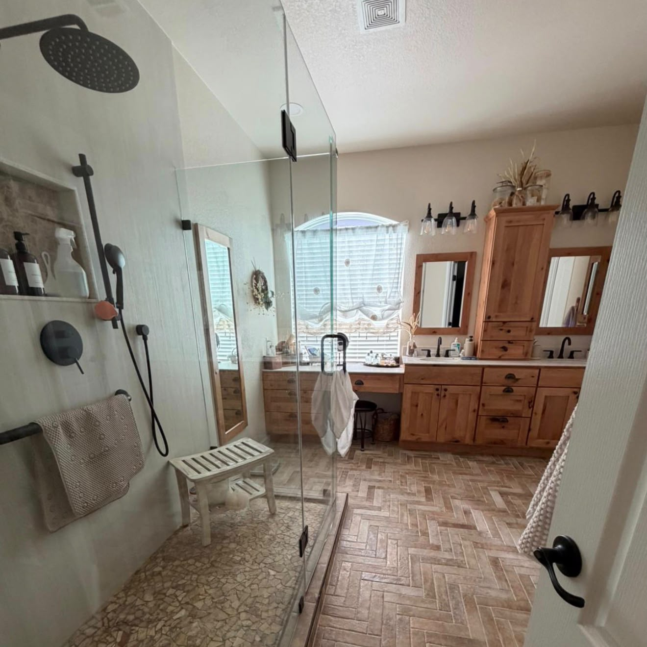 Glass-enclosed shower with pale walls and black fixtures, with wood-toned cabinetry and framed mirrors beyond.